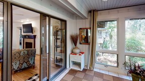 A modern living room with a sofa, a television, a decorative vase, and large windows offering a view of greenery. - Exquisitely Furnished Home Surrounded by Golf Courses, Tennis Courts, & Beaches (Kiawah Island)