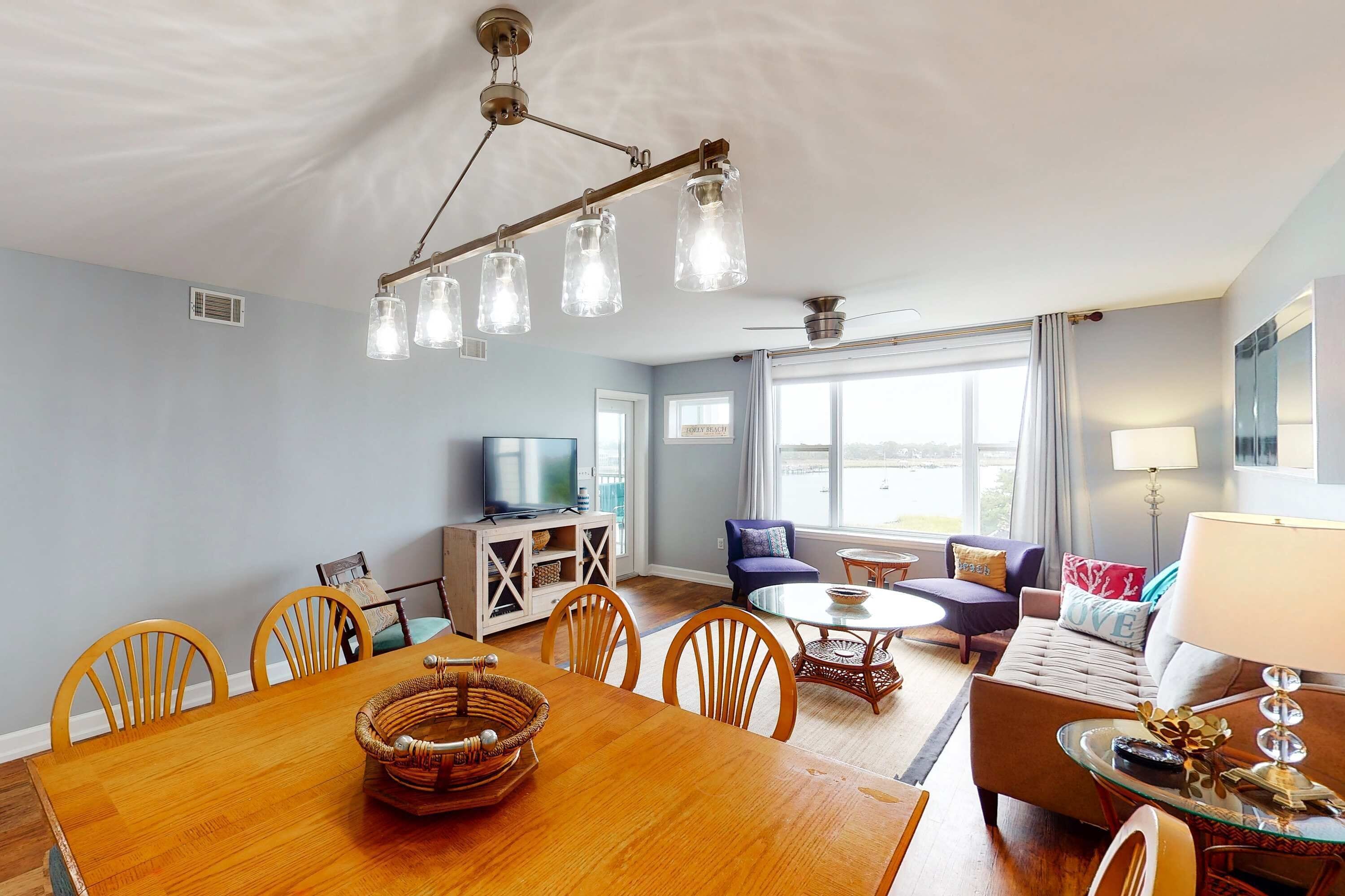 A spacious living room with a wooden dining table, comfortable seating, a television, and large windows.