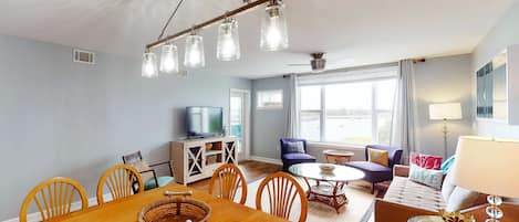 A spacious living room with a wooden dining table, comfortable seating, a television, and large windows.