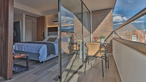 A modern bedroom with a large bed, a small table, and a balcony with wicker chairs and a potted plant. - Edificio Bloom (Medellín)