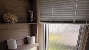 A modern bathroom with a toilet, a window with blinds, a shelf with toilet paper, and a small decorative item. - 3 Bedroom Caravan Alix (Rhyl)