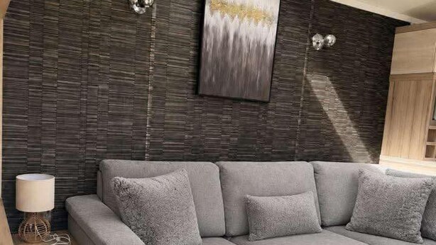 A modern living room with a grey sectional sofa, a wooden side table, a floor lamp, and a large abstract painting on the wall. - 3 Bedroom Caravan Alix (Rhyl)