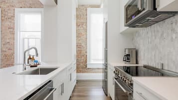 Une cuisine moderne avec des meubles blancs, un mur d’accent en briques, des appareils électroménagers en acier inoxydable et une grande fenêtre.