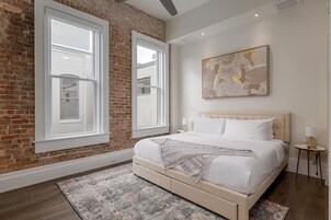 Un dormitorio con pared de ladrillo, ventanas amplias, cama con ropa blanca y un cuadro abstracto moderno.