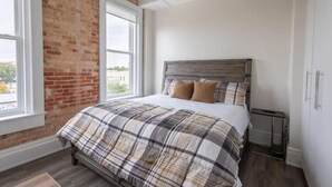 A bedroom with a bed, a window, and a brick wall. - The Brent Lofts (Pensacola)