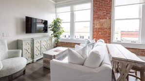 A modern living room with a white sofa, a wooden table, and a large window with a brick wall. - The Brent Lofts (Pensacola)