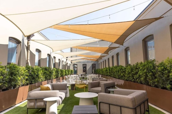 Un área de lounge al aire libre moderna, con asientos y mesas, rodeada de vegetación y protegida por grandes toldos.
