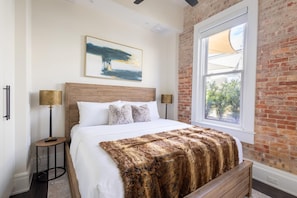 Un dormitorio con una cama grande, cabecera de madera, ventana con vistas, un cuadro en la pared y una lámpara en una mesita de noche.