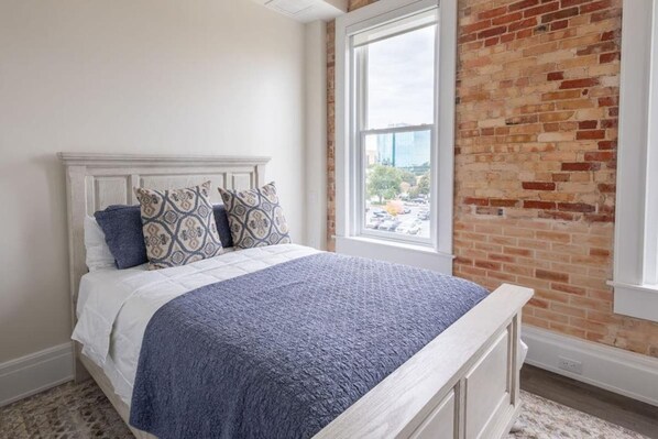Un dormitorio con una cama blanca, almohadas estampadas, una colcha azul, un ventanal grande y una pared de ladrillo.