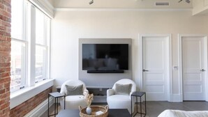 Een moderne woonkamer met een flatscreen televisie aan de muur, twee witte fauteuils, een zwarte salontafel en een bakstenen muur.