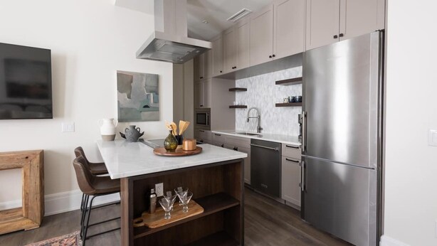 A modern kitchen with a stainless steel refrigerator, a central island, and a dining area with chairs. - The Brent Lofts (Pensacola)