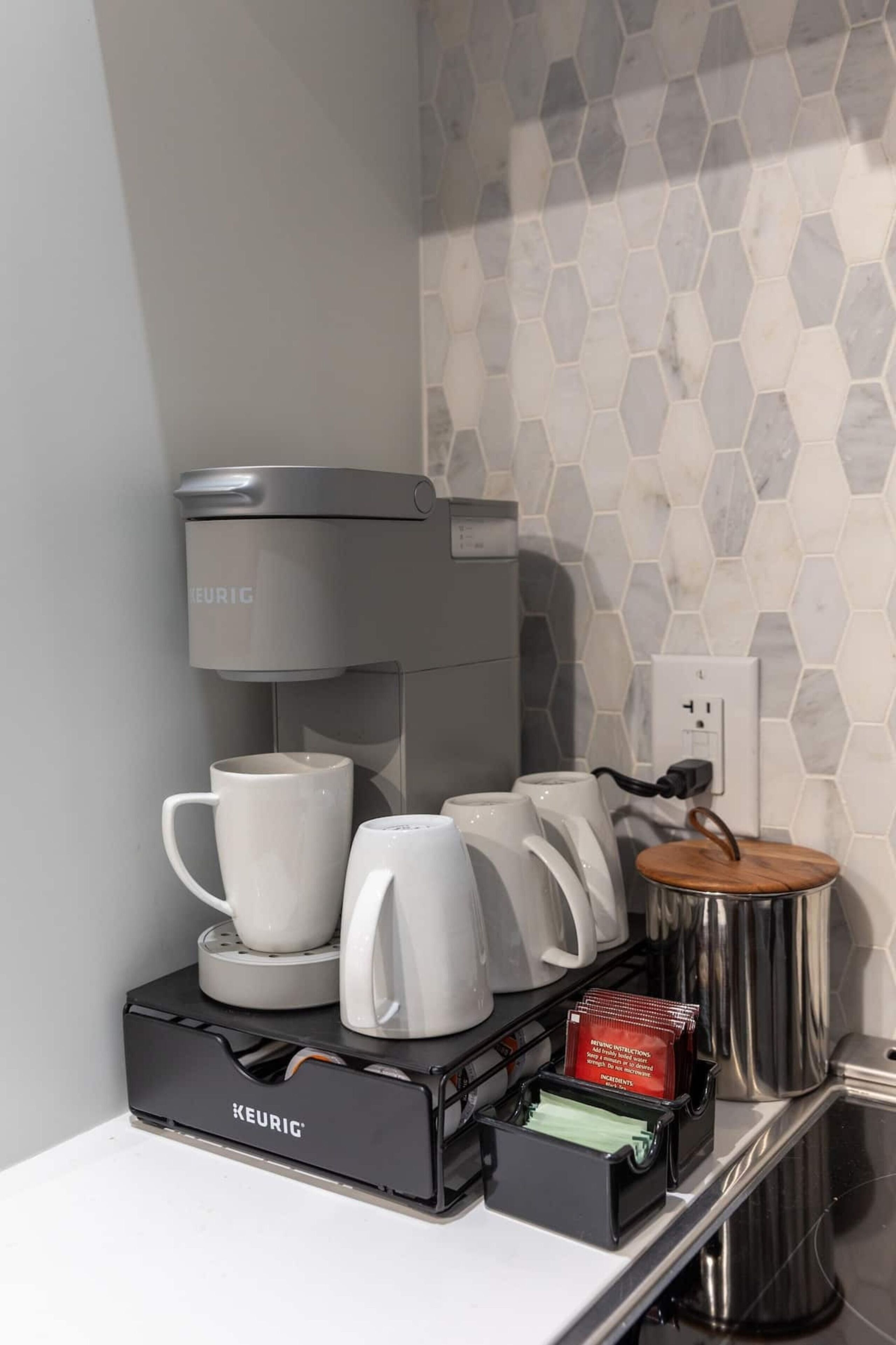 Eine KEURIG Kaffeemaschine steht auf einer Arbeitsplatte mit zwei weißen Tassen, einer Kaffeekanne und einem Mülleimer.