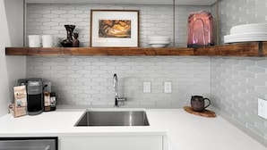 A modern kitchen with white cabinets, a hexagonal tile backsplash, a stainless steel sink, and a coffee maker. - The Brent Lofts (Pensacola)