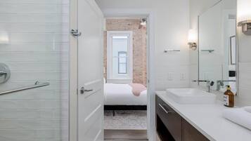 A modern bathroom with a shower, a large mirror, and a bed in the background.