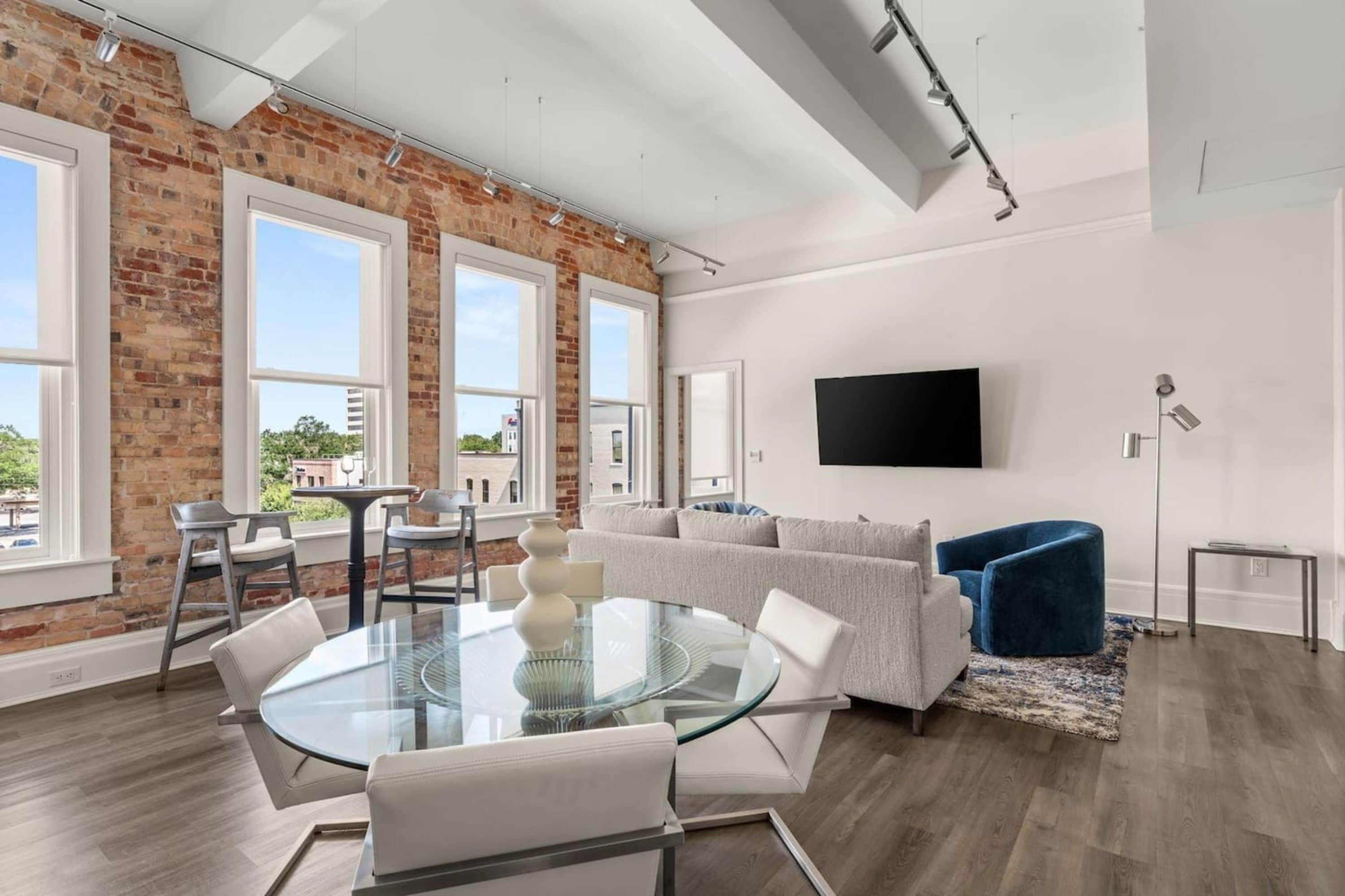 A modern living room with a glass table, white sofa, and blue chair, a flat-screen TV, and large windows with a brick wall.