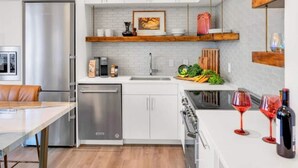Een moderne keuken met witte kasten, een roestvrijstalen koelkast, een vaatwasser en een fornuis. Daarboven bevindt zich een houten plank boven de gootsteen, met decoratieve objecten en een lijst met een foto.