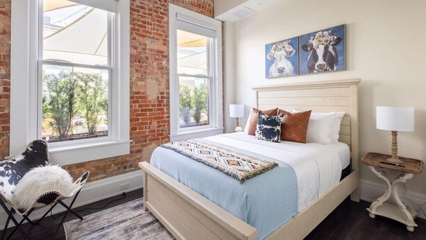 Een slaapkamer met een bakstenen muur, twee ramen, een bed met hoofdeinde, een stoel en kunst aan de muur.