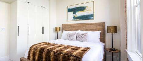 A bedroom with a wooden bed, white walls, a large painting, and a nightstand with a lamp.
