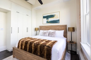 Una camera da letto con un letto in legno, pareti bianche, un quadro di grandi dimensioni e un comodino con una lampada.