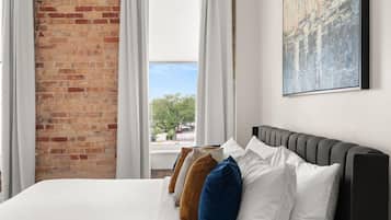 A hotel room with a brick wall, a bed with white and colored pillows, a painting on the wall, and a window with curtains.