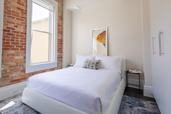 Un dormitorio con pared de ladrillo, un ventanal grande, una cama con ropa de cama blanca, una mesita de noche y una obra de arte enmarcada en la pared.