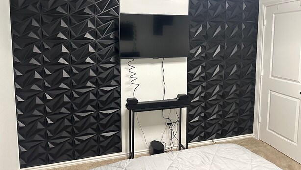 A room with a black geometric-patterned wall, a mounted flat-screen TV, a small shelf with a plant, and a bed with white bedding. - Cozy Hideaway with Pool – Relax & Unwind Just Off the Highway (Maxwell)