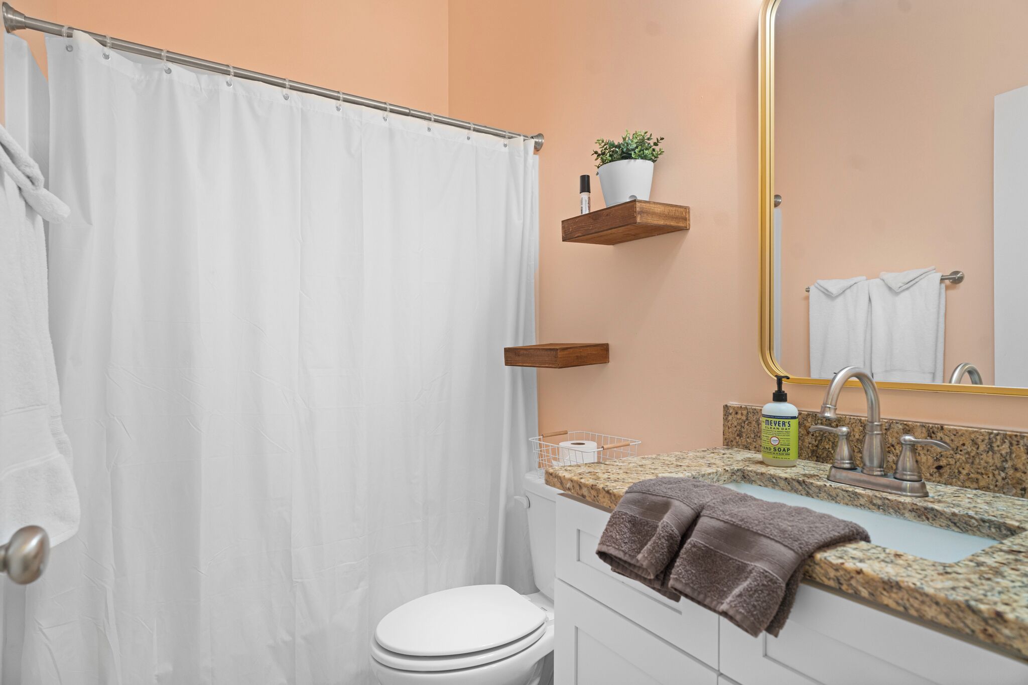 Une salle de bain avec des toilettes blanches, un rideau de douche, un lavabo avec un plan de travail en granit, un miroir et un porte-serviettes.