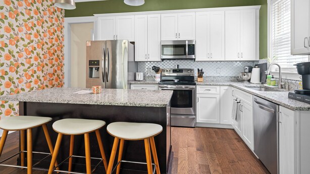 A kitchen with white cabinets, a granite countertop, stainless steel appliances, and wooden stools. - The Peach House- Near Downtown (Savannah)