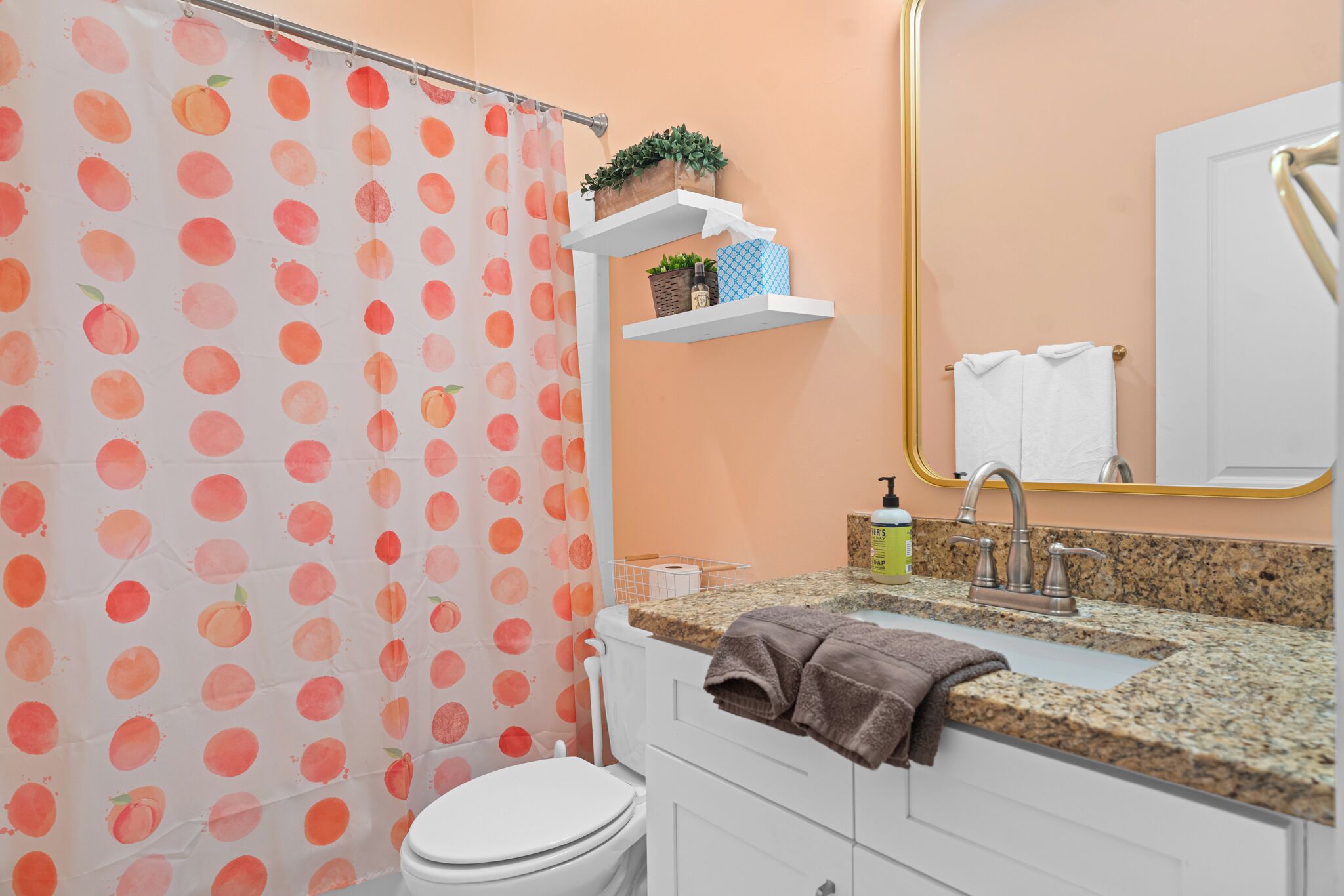 A bathroom with a peach-colored shower curtain, a white toilet, a sink with a granite countertop, and a mirror with a gold frame.