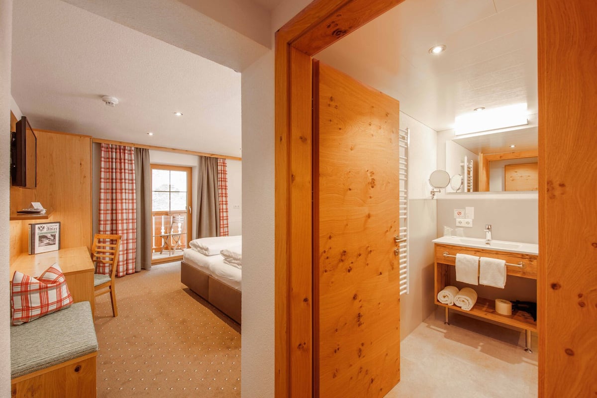 A hotel room with a bed, a wooden bench, a small table with a picture frame, a window with curtains, and a bathroom with a sink and mirror.