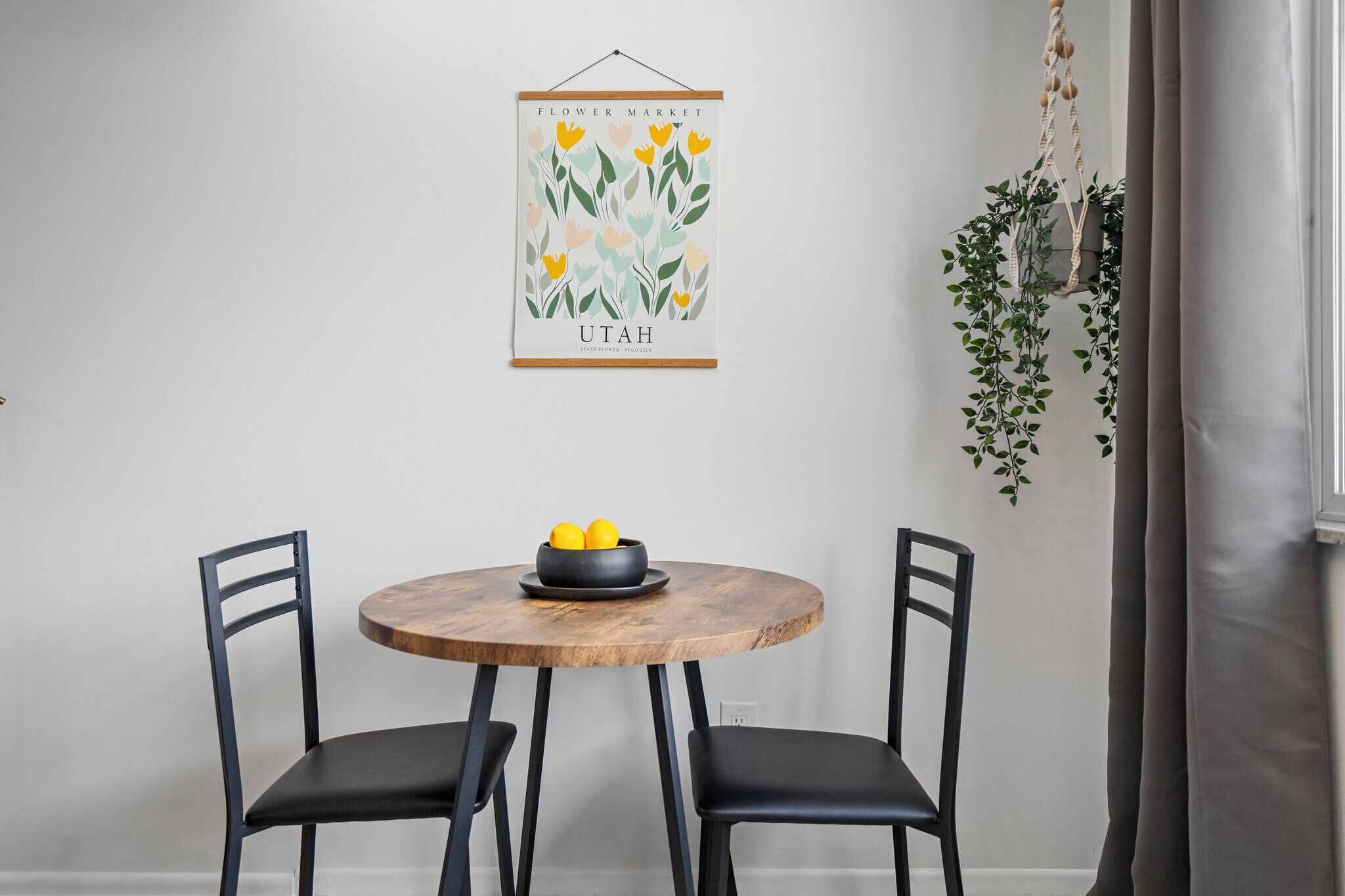 A dining area with a round wooden table, two black chairs, and a hanging macrame plant.