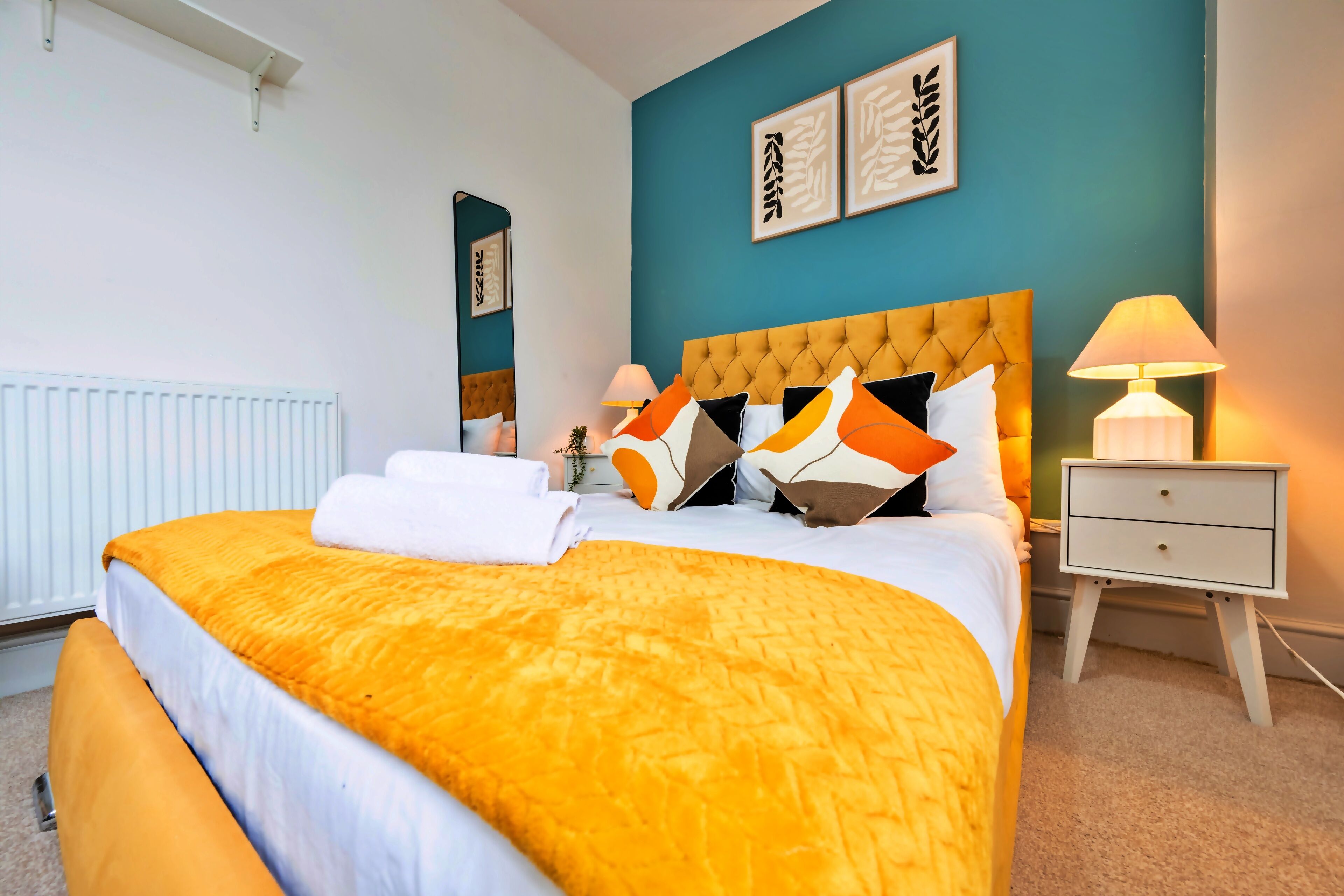 Una cama bien hecha con cabecera amarilla, ropa de cama blanca y almohadas naranjas. Hay una mesita de noche con una lámpara y un cajón pequeño. La habitación tiene paredes color esmeralda y un radiador blanco.