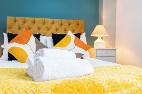 A neatly made bed with a yellow quilt, white pillows with orange and black accents, and a folded white towel on the bed. - The Ground Floor Getaway (Cardiff)