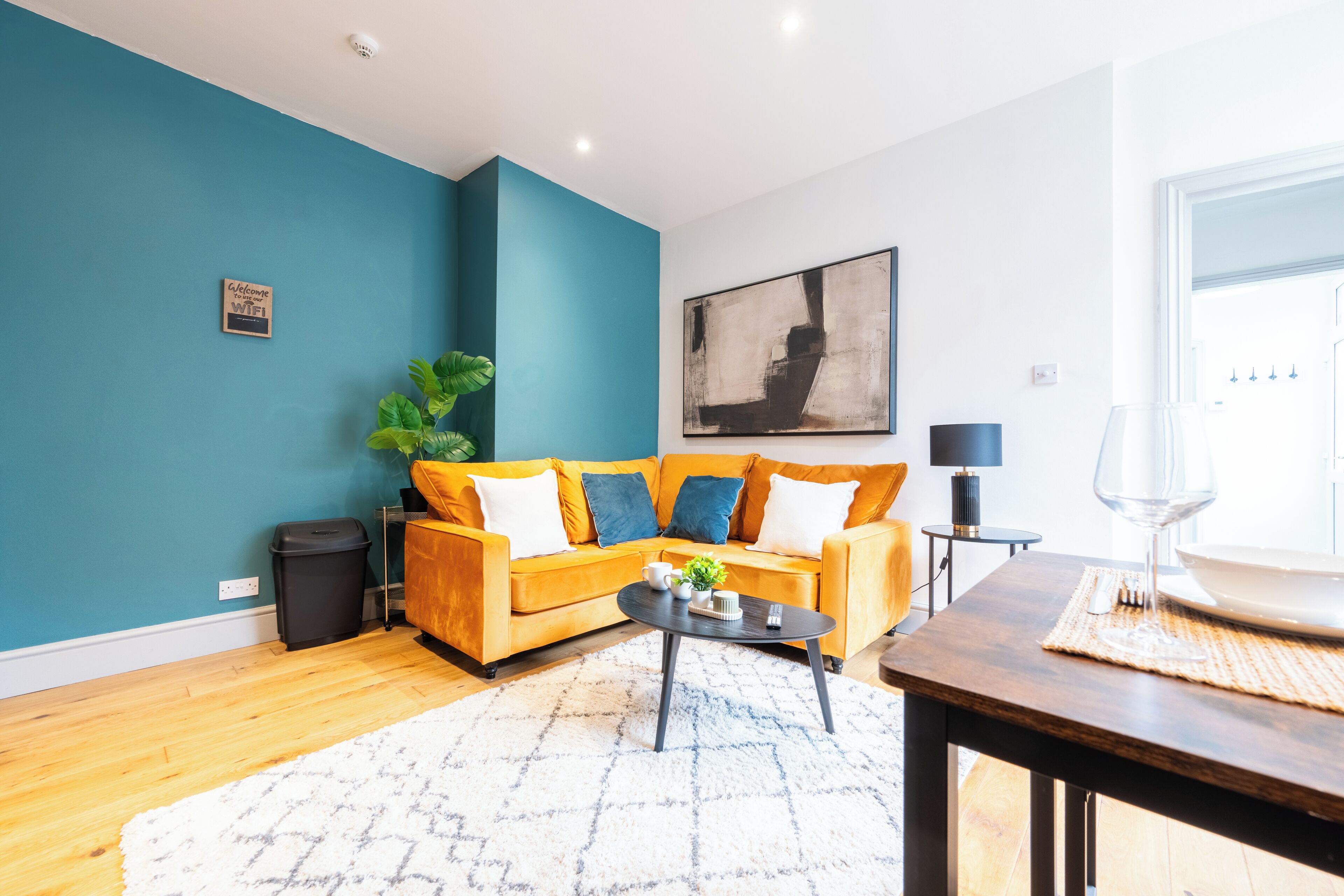 A living room with a teal wall, a yellow sofa, a coffee table, and a dining area with a glass of wine.