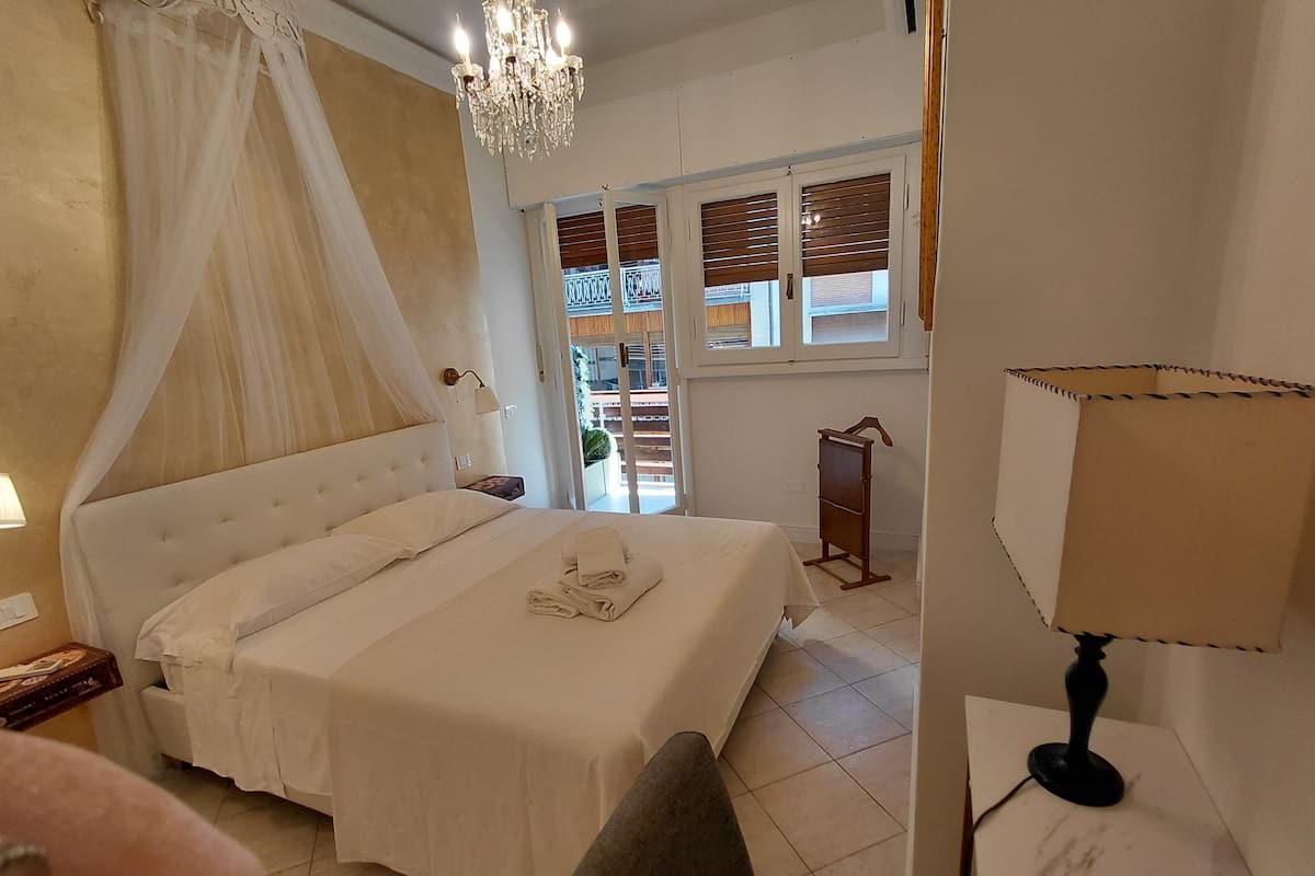 A neatly made bed with a canopy, a bedside table with a lamp, and a view of the outside balcony through the window.