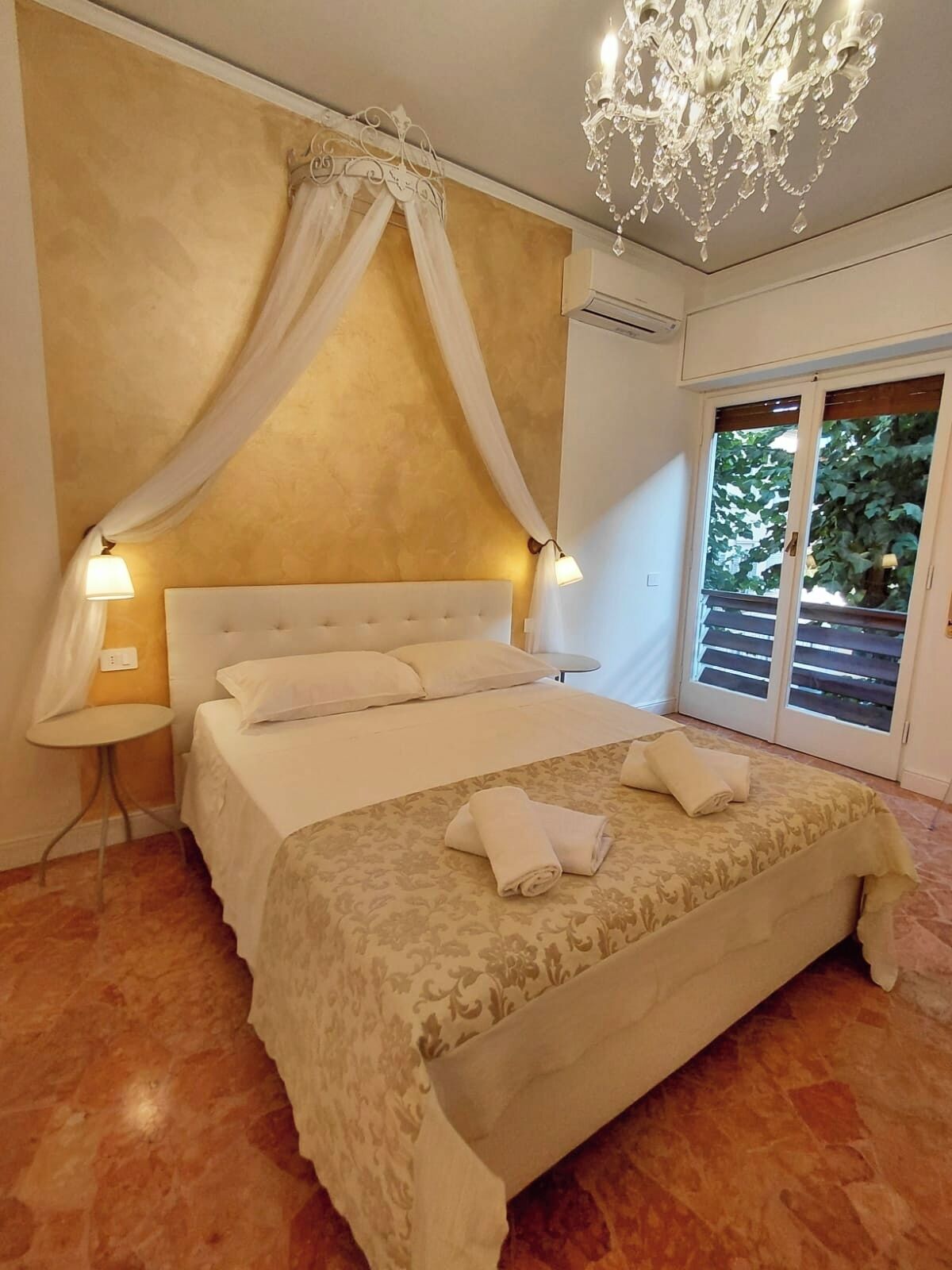 Een slaapkamer met een groot bed, een kroonluchter, een klein tafeltje en een raam met uitzicht op groen.