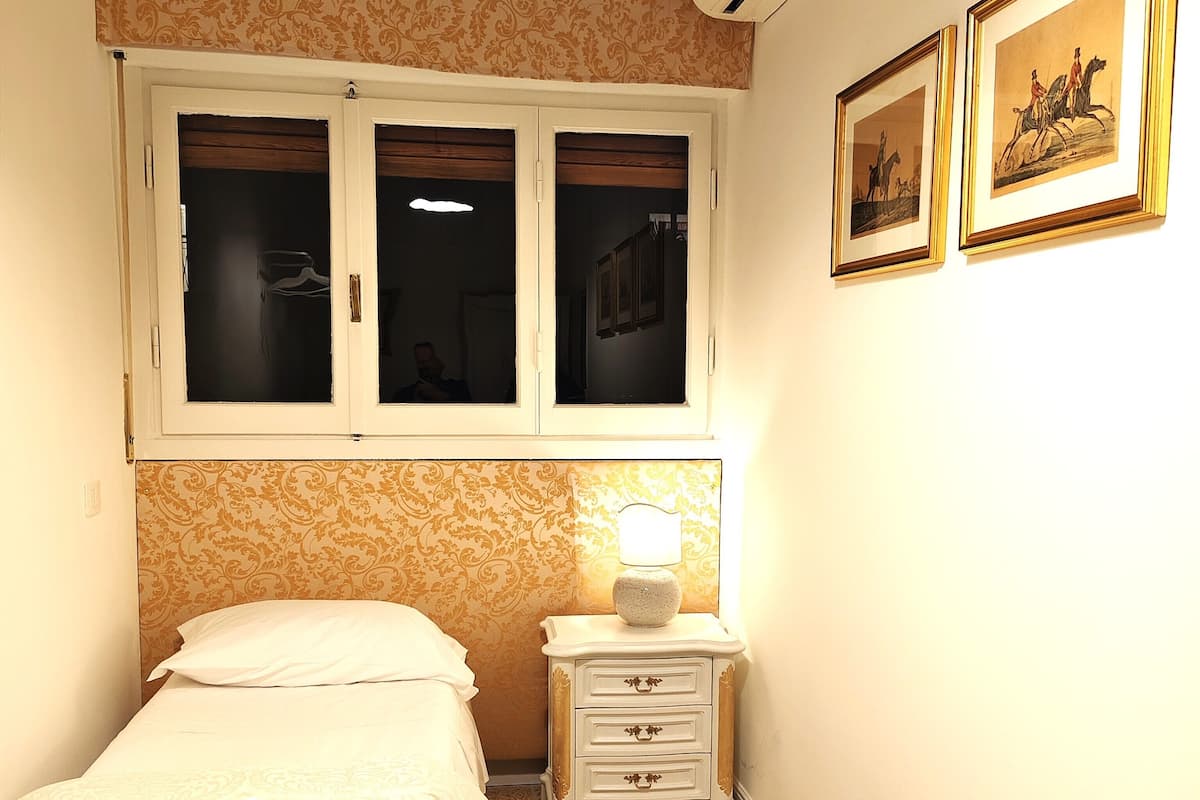 A bedroom with a bed, a nightstand with a lamp, a window with curtains, and two framed pictures on the wall.