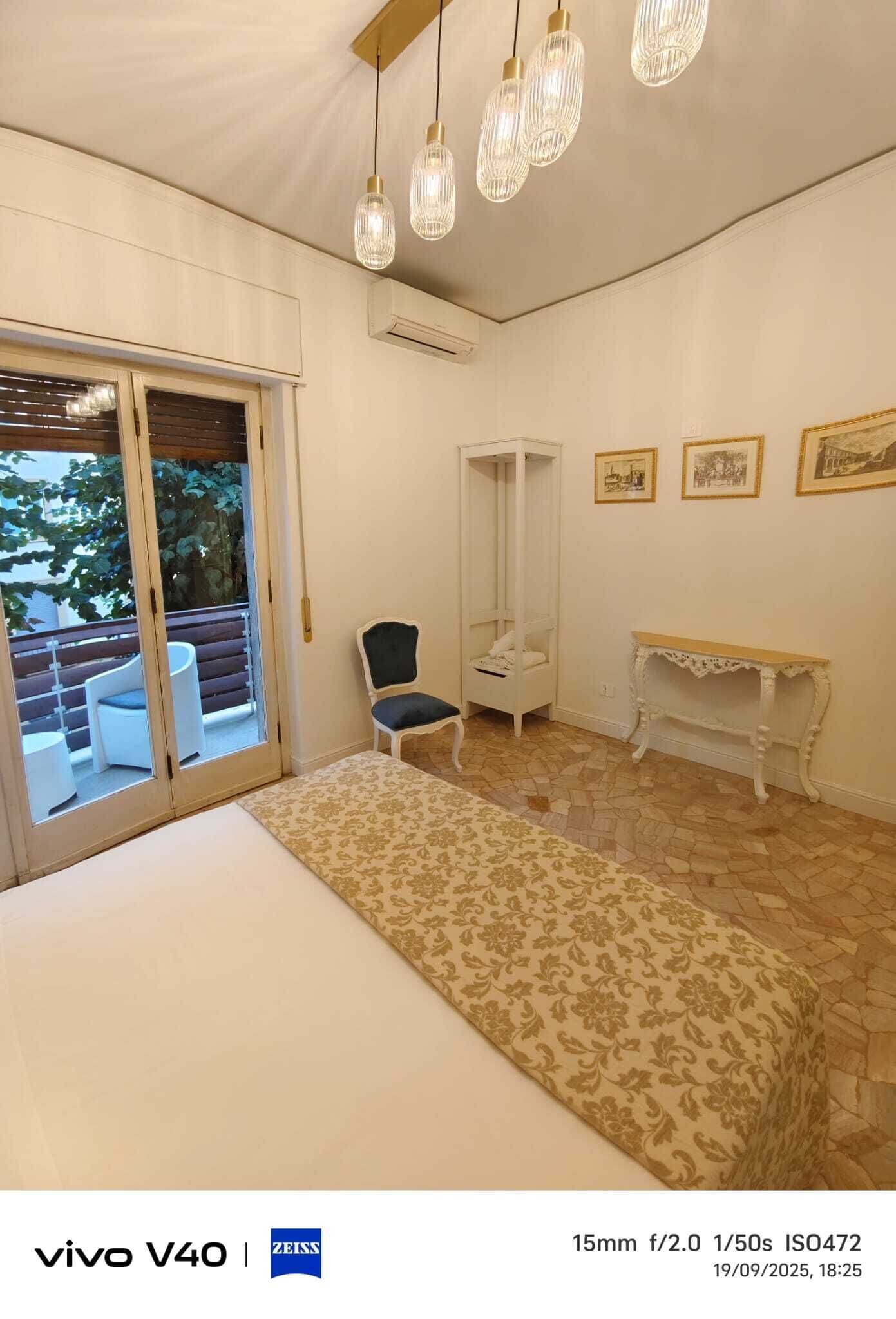 A room with a bed, a chair, a small table, and a wardrobe. There is a sliding door leading to an outdoor area with a bench and a table.