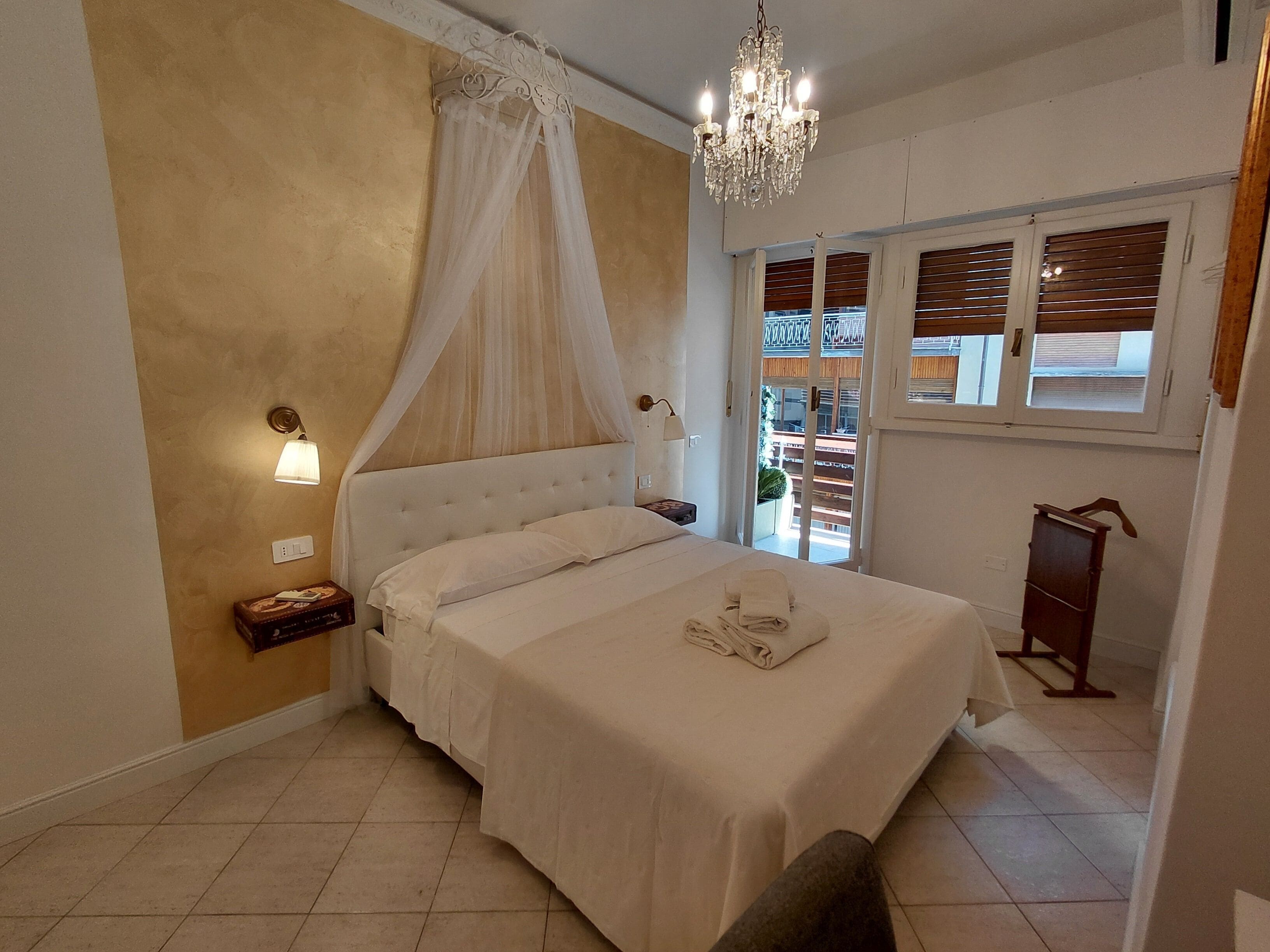 A bedroom with a canopy bed, a chandelier, a nightstand, and a view of the outside balcony.