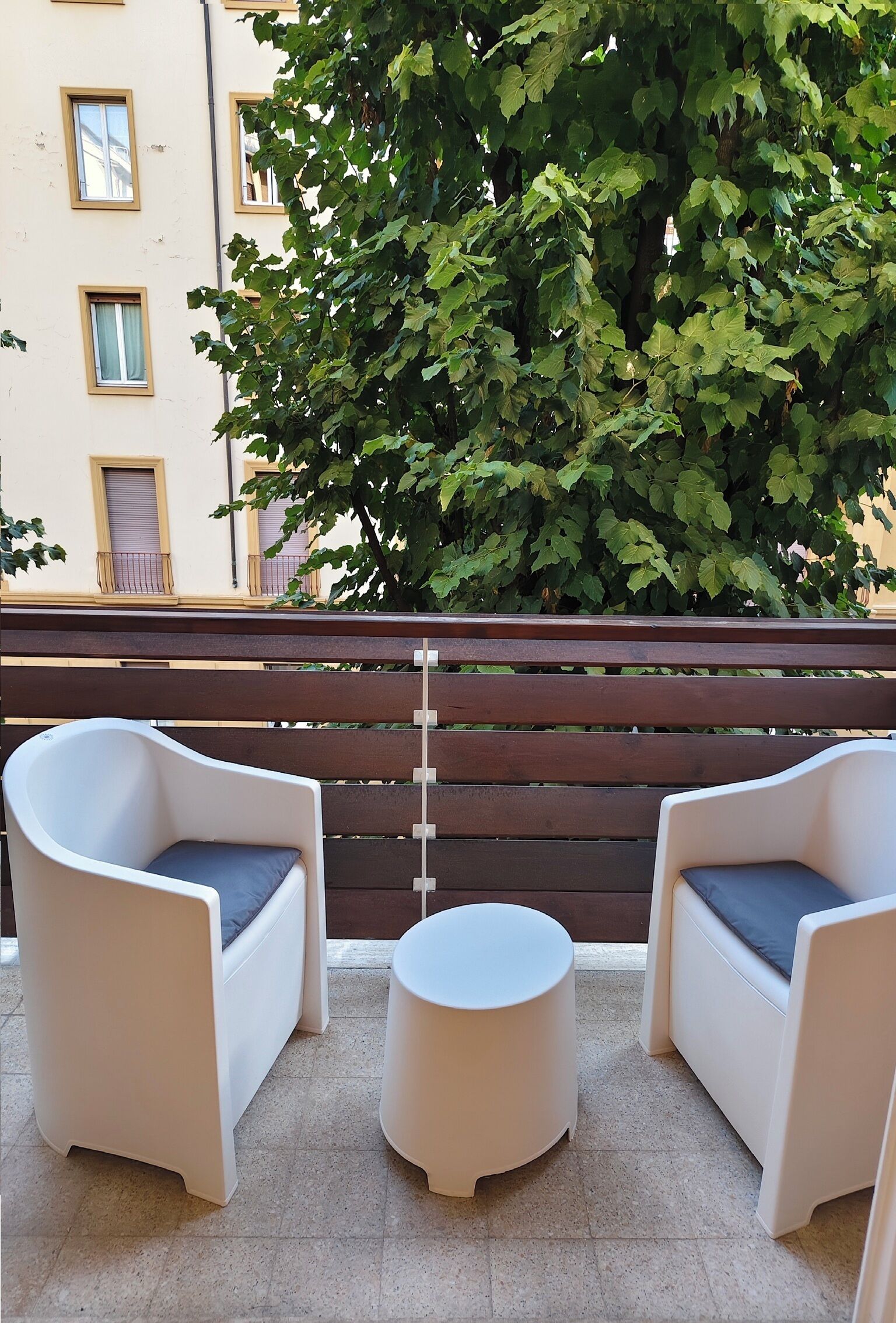 Ein Balkon mit zwei weißen Sesseln, einem kleinen runden Tisch, einem Holzzaun und einem großen Baum mit grünen Blättern.