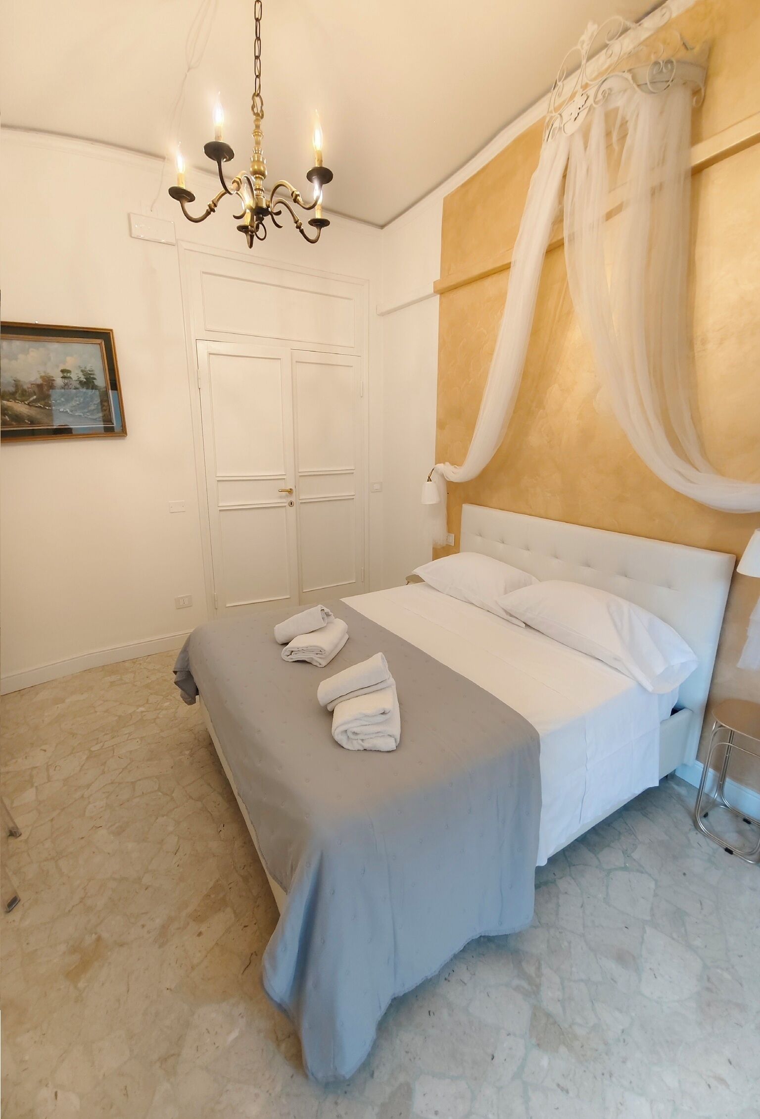 Een slaapkamer met een bed, witte gordijnen, een kroonluchter en een schilderij aan de muur.