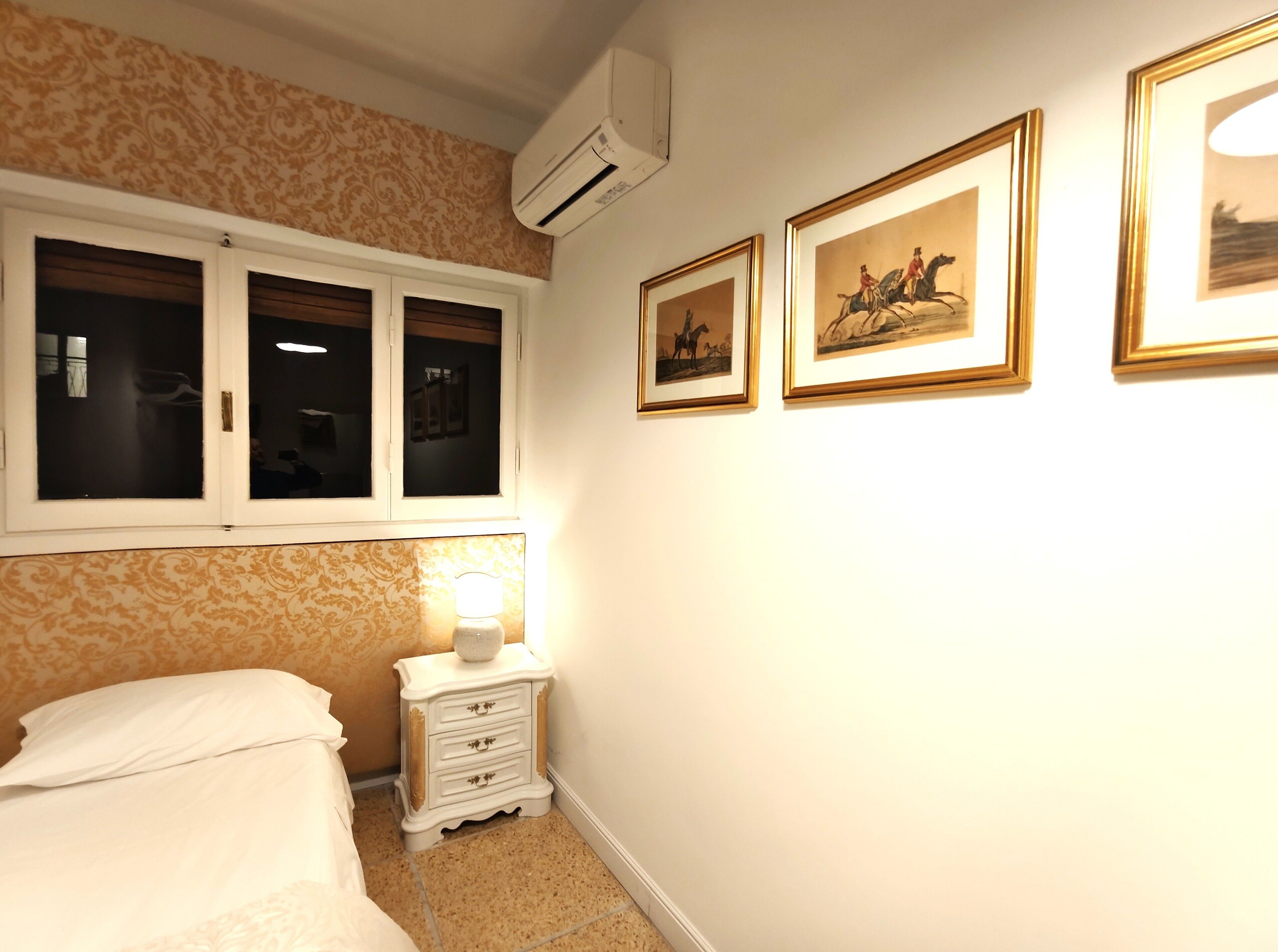 A bedroom with a bed, a nightstand with a lamp, a window with closed shutters, and three framed pictures on the wall.