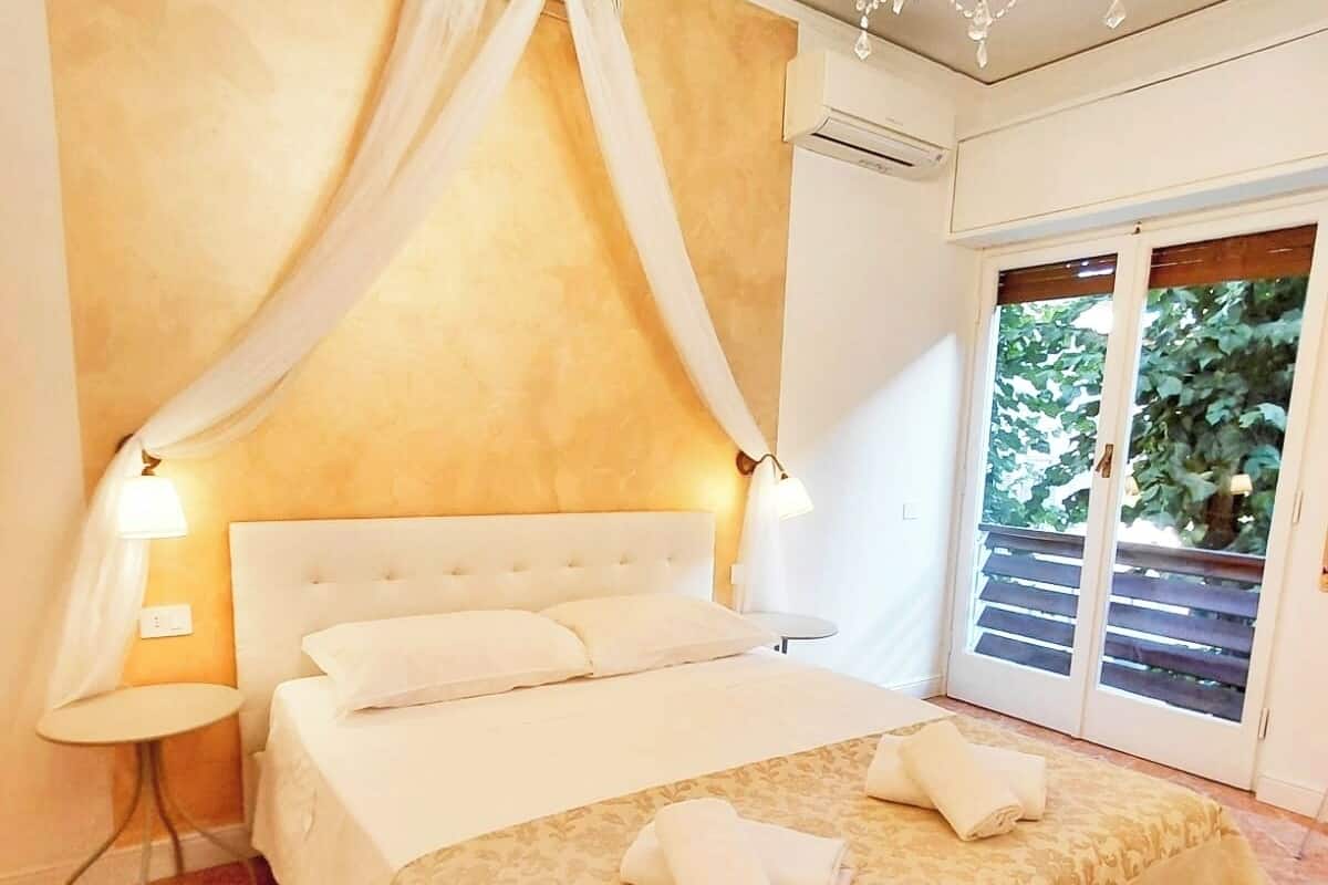 A bedroom with a large bed, a canopy, a chandelier, and a view of greenery through the window.