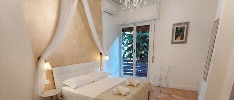 A bedroom with a bed, a chandelier, a window with a view of greenery, and a clear chair.