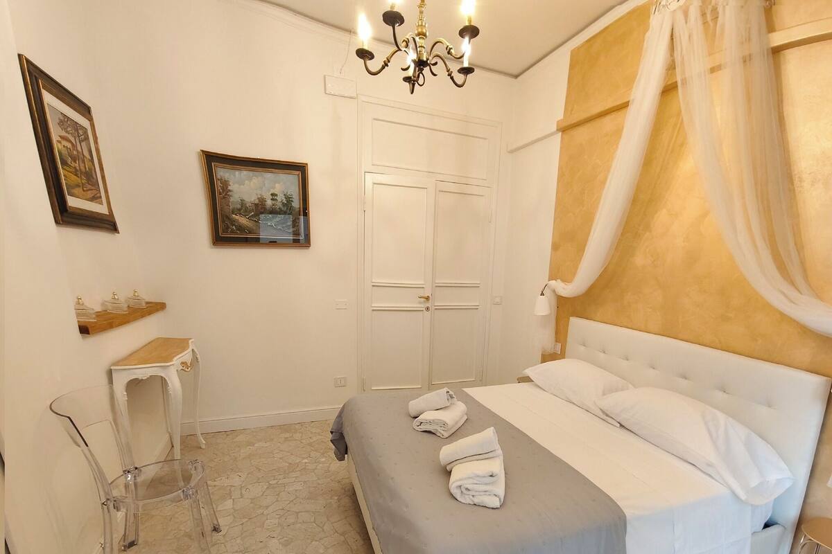 A bedroom with a bed, a nightstand, a chair, a small table, and a chandelier.