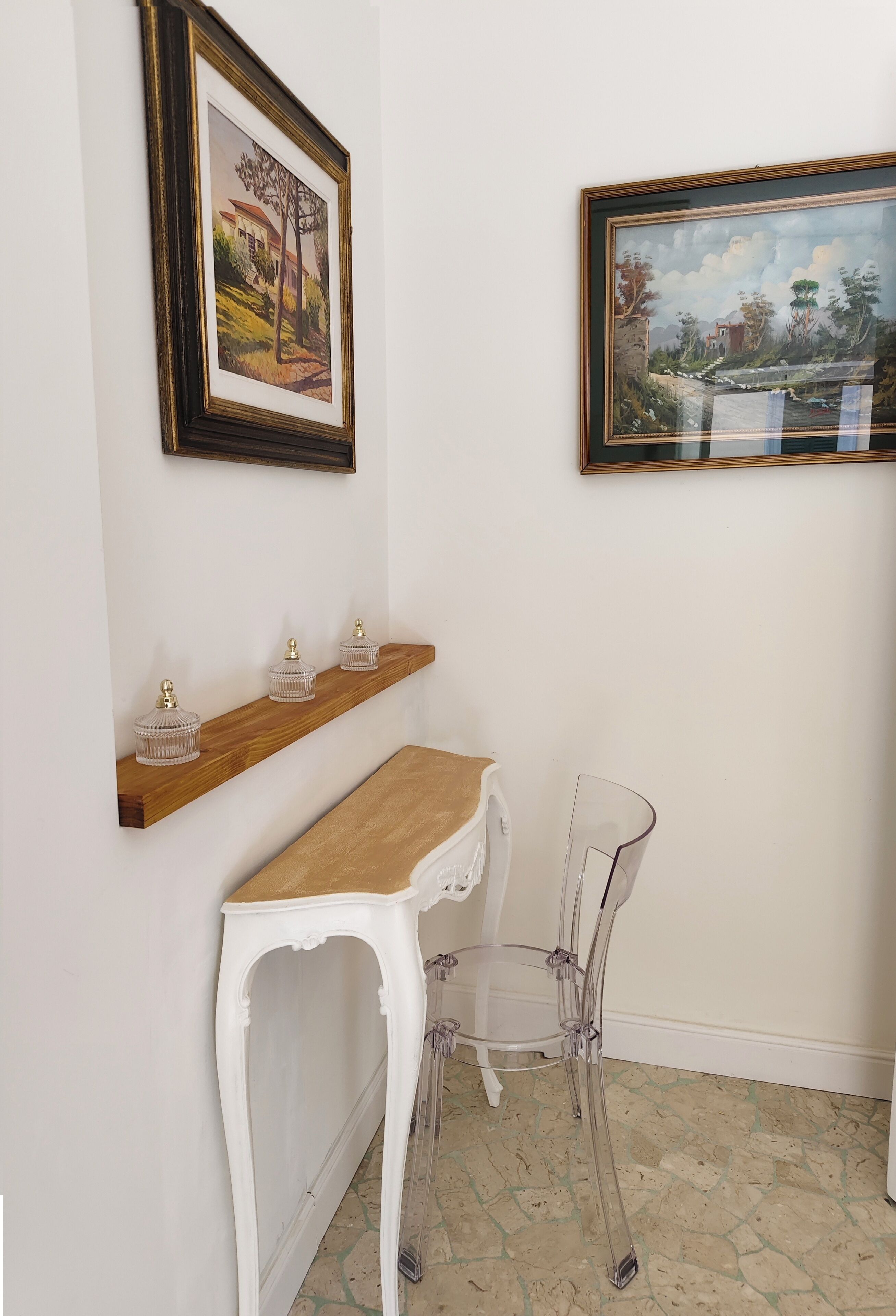 A room with a wooden table, two chairs, a shelf with decorative items, and two framed paintings on the wall.