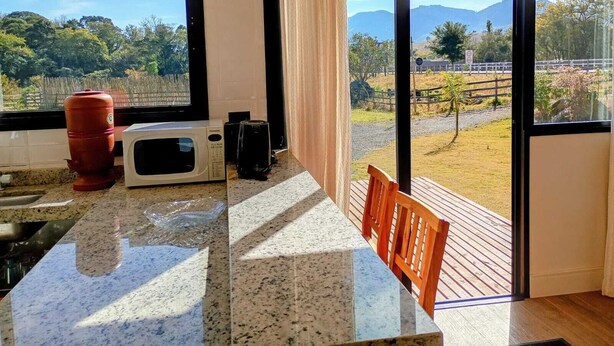 Une cuisine avec une grande fenêtre, un four à micro-ondes, un distributeur d’eau rouge et une chaise en bois.