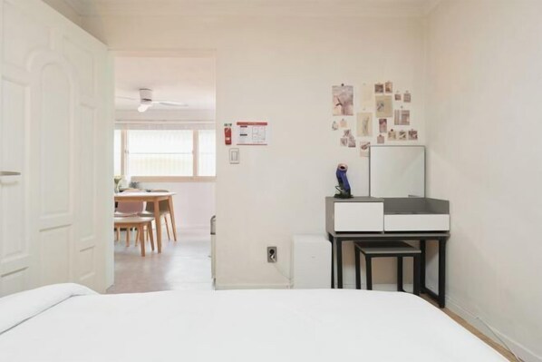 A bedroom with a bed, a desk with a mirror, and a small table. There is a door leading to another room, and a window with blinds.