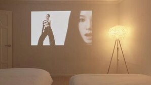 A bedroom with a bed, a wall-mounted projector displaying a person, and a floor lamp.