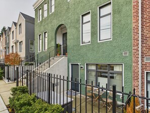 Eine Reihe von Reihenhäusern mit grüner, strukturierter Fassade, einem Hauseingang mit Treppe und einer kleinen Terrasse mit Gartenmöbeln.